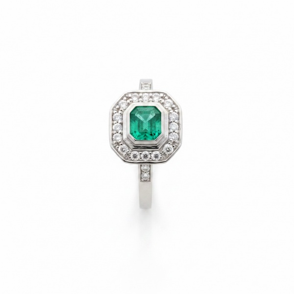 Anel em ouro branco 18k, peso aproximado de 4 g. Apresenta esmeralda natural em lapidação retangular (emerald cut), com peso estimado de 0,60 ct, engastada em bezel, circundada por halo de brilhantes