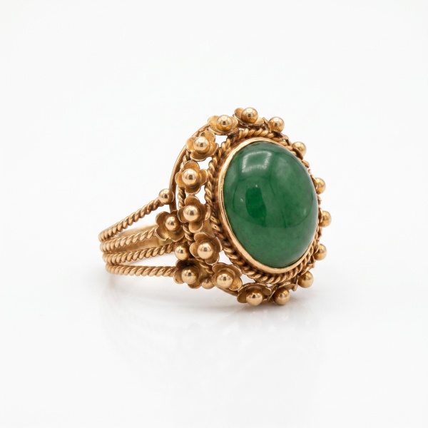 Anel em ouro amarelo com cabochão oval de pedra verde ao centro, moldura em cordão trançado e bordadura ornamental com esferas e floretes em ouro. Design vintage.Peso: 6,8 g | Aro: 16