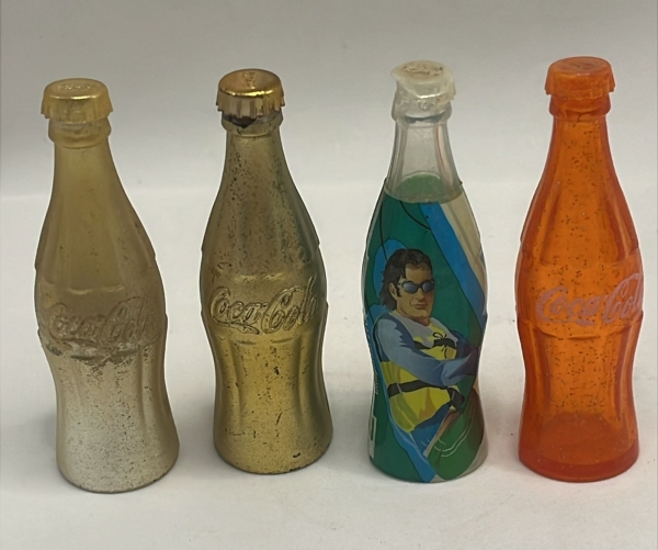 Lote com quatro miniaturas de garrafas Coca-Cola colecionáveis, incluindo dois modelos metalizados em tom dourado, uma versão temática com ilustração esportiva e uma peça em plástico translúcido laran