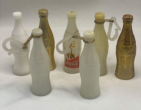 Lote com sete miniaturas colecionáveis de garrafas Coca-Cola, incluindo modelos em plástico branco, versões metalizadas douradas e peças equipadas com alças plásticas de suporte. Conjunto vintage dive