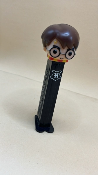 Porta-balas PEZ colecionável do personagem Harry Potter, identificado pela cicatriz de raio e o brasão da escola Hogwarts na haste preta. O item possui aproximadamente 11 cm de altura
