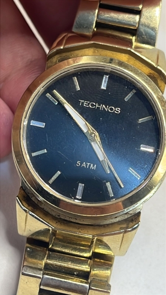 Relógio Technos dourado, modelo 2035.MCM, apresenta um design clássico com mostrador preto e marcadores em relevo. A peça possui caixa de aproximadamente 38 mm (incluindo a coroa), resistência de 5 AT