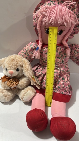 Coelho de Pelúcia Bege Sentado com Laço Marrom no Pescoço, Segurando Pequena Cesta de Palha Trançada, Acompanhado de Boneca de Pano com Vestido Florido Rosa. Aproximadamente
