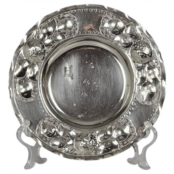 Salva de formato circular, de metal prateado. Apresenta espelho central liso e plano com leve rebaixamento. Destaca-se pela aba larga ricamente ornamentada em repuxado e cinzelado, exibindo profusa de