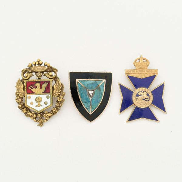 Conjunto contendo tres broches comenda em metal dourado, a saber: 1- Brasão de armas, produzido pela famosa marca de joias norte americana "Coro", apresentando escudo dividido em duas seções principai