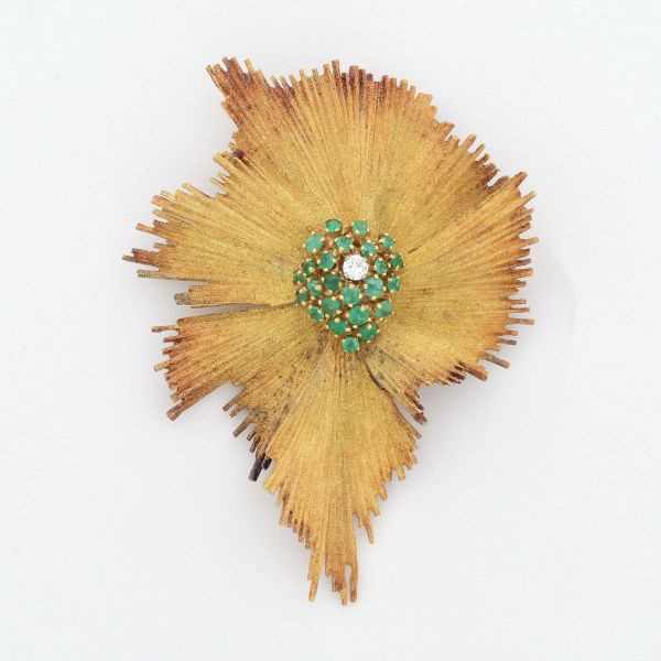 Broche em ouro amarelo 18 k, em formato de flor, caracterizado com pétalas desfiadas ou texturizadas, tendo ao centro pavê de pequenas esmeraldas e um diamante central de aproximadamente 0,10 ct. Cont