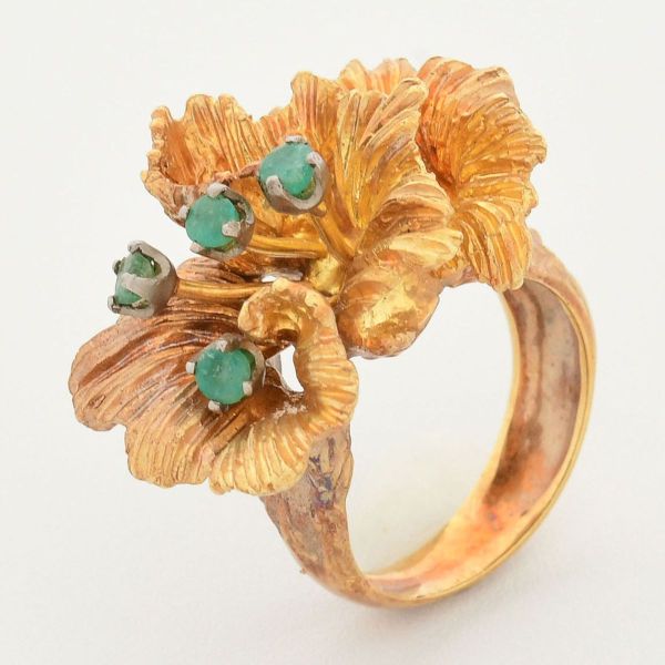 Anel em ouro amarelo 18 k, em formato de flor, caracterizado com pétalas desfiadas ou texturizadas, tendo ao centro, quatro pequenas esmeraldas. Aro 13. Peso total 14,4 g.