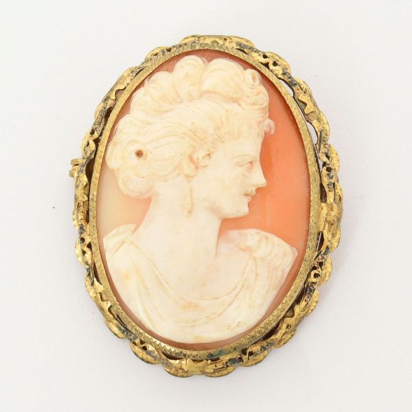 Broche pendente camafeu, representando figura feminina em relevo claro com fundo de tom alaranjado, sobre concha natural, emoldurado em dourado. Medida 53 x 43 mm.
