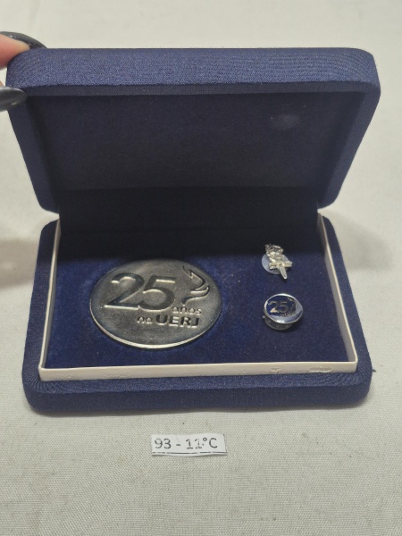 Estojo comemorativo dos 25 anos na UERJ, com uma medalha e 2 botons. Medindo a medalha 5,5cm de diâmetro.