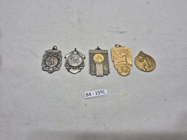 Conjunto de 5 medalhas e insígnias esportivas em metal, incluindo peças com os anéis olímpicos, representações de voleibol, remo e futebol, apresentando acabamentos em tons de prata e ouro com relevos