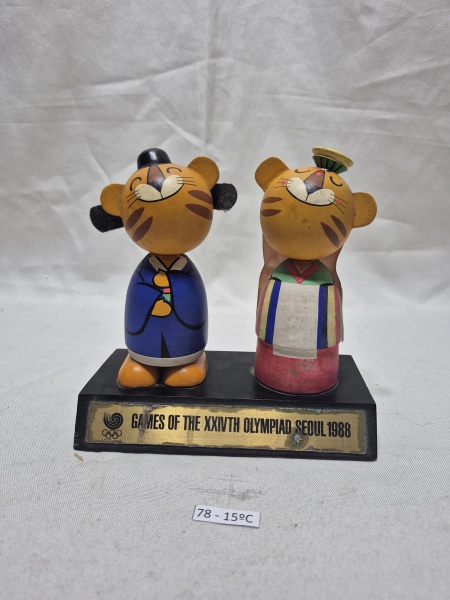 Conjunto comemorativo oficial das Olimpíadas de Seul 1988, apresentando o mascote Hodori e sua contraparte feminina em figuras sobre base de madeira com placa identificativa. Medindo 15cm x 18cm x 14c