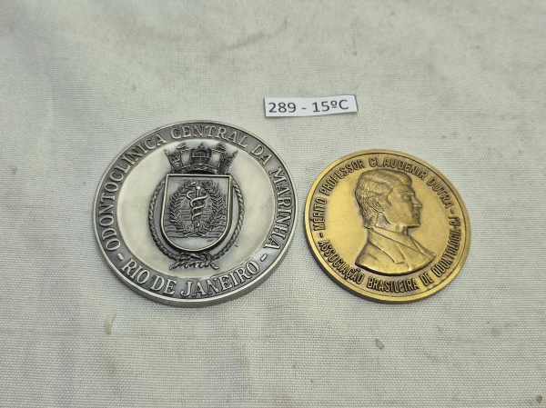 Conjunto de 2 medalhas de odontologia, sendo 1 grande em metal prateado comemorativa de 50 anos da Odontoclínica Central da Marinha (1935-1985) conferida ao Dr. C. A. Pego e 1 de Mérito Professor Clau