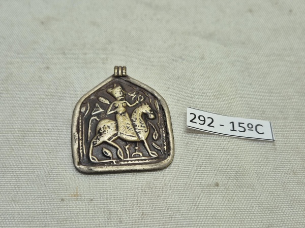 Pingente em prata de formato irregular com passador, apresentando figura em relevo de divindade ou figura histórica montada em cavalo. Medindo 4cm x 3,5cm.