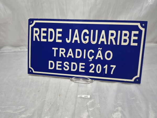 Placa em plástico duro decorativa em tom azul com inscrições em branco "Rede Jaguaribe - Tradição Desde 2017", com furações nos cantos para fixação. Medindo 40cm x 20cm.