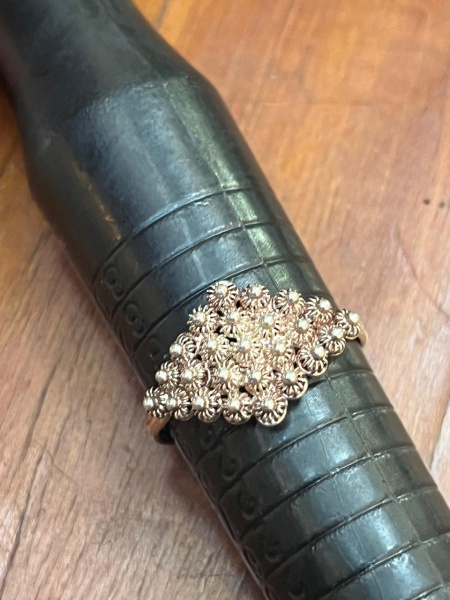 Anel de estilo chuveiro ou filigrana vintage, caracterizado por uma cabeça proeminente em formato de buquê ou padrão floral detalhado feito de esferas e texturas metálicas.