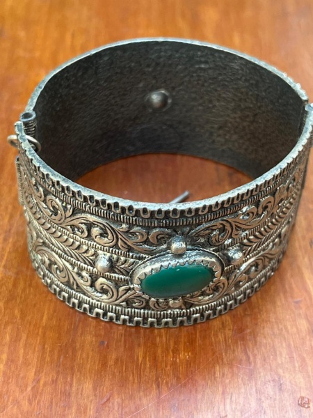 Bracelete vintage de metal com design étnico, caracterizado por ricos detalhes gravados em relevo arabesco e texturas envelhecidas. A peça traz uma pedra cabochão central oval na cor verde, que simula