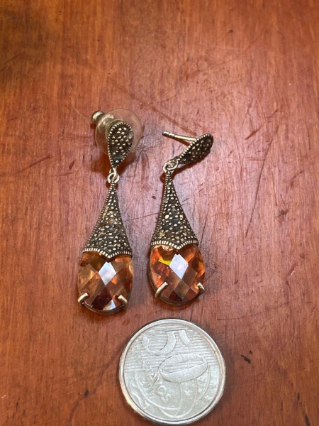 Brincos são joias em estilo vintage, caracterizados por uma base detalhada com pequenas pedras de marcassita e um pingente facetado em formato de gota em cristal de cor champanhe ou citrino.