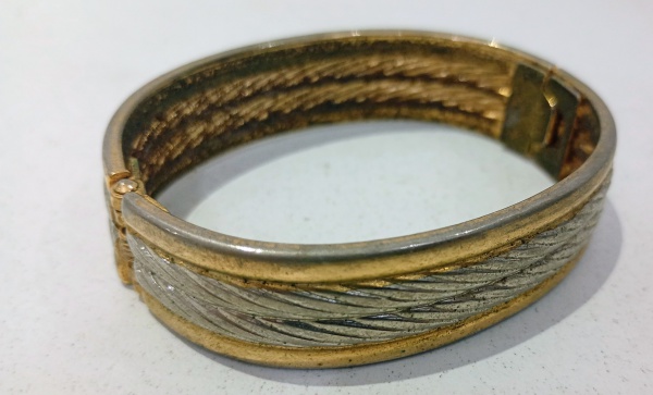 Pulseira estilo bracelete rígido articulado caracterizada pelo seu design bicolor que combina tons dourados nas bordas e prateados na parte central. A faixa central apresenta um padrão texturizado dia
