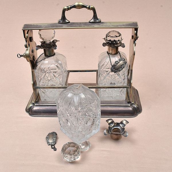 Tântalus inglês composto por elegante suporte retangular com alça superior e mecanismo de travamento lateral prateado abrigando três garrafas em cristal lapidado, de formato abaulado e ombros arredond