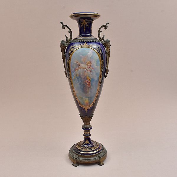 Ânfora em porcelana francesa de Sèvres do sec. XIX. Corpo alongado com gargalo afunilado e boca evasê, guarnecida por alças em bronze patinado no formato de volutas estilizadas, com máscaras/bustos fe