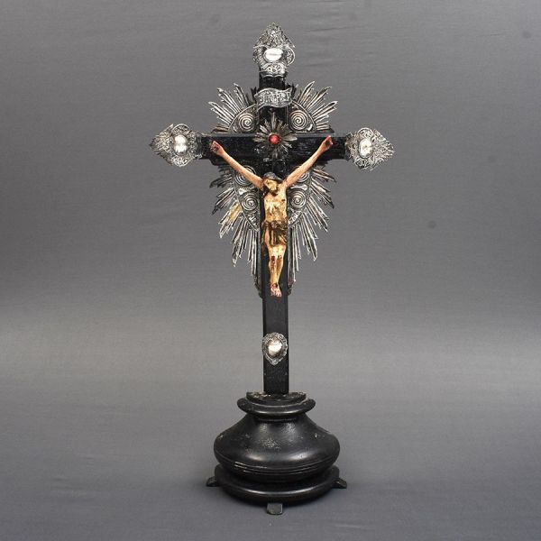 Crucifixo sul americano com cruz de madeira de linhas retas, montada sobre peanha circular. Nos arremates dos três braços superiores e no cruzeiro, aplicações de metal prateado cinzelado, com motivos