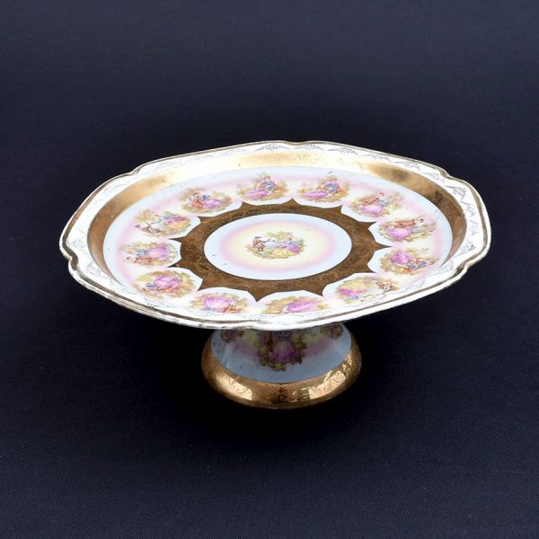 Fruteira/prato de pé alto (compotier) de porcelana francesa, decorada em policromia e dourado no gosto Louis XV, com cenas galantes de inspiração dieciochesca. Ao centro medalhão com casal em cena rom