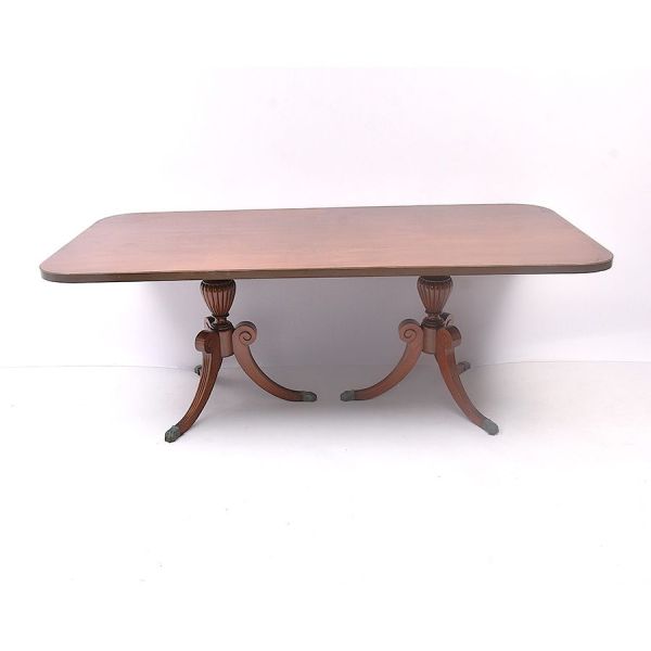 Mesa de jantar inglesa em madeira nobre estilo Regency, de duplo pedestal (`twin-pedestal table`). Tampo de perfil ovalado, com friso em metal aplicado à borda, apoiado sobre dois suportes centrais em