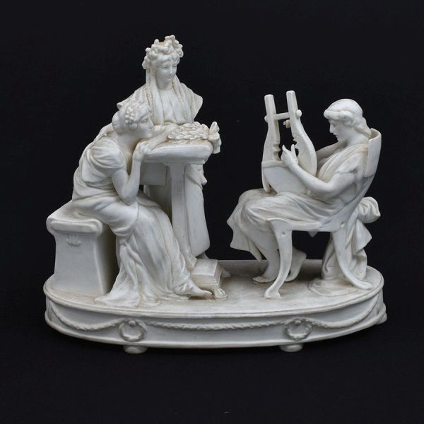 Grupo escultórico francês em biscuit branco, representando refinada cena de inspiração neoclássica, com três figuras femininas trajadas à maneira da Antiguidade Clássica reunidas em torno de uma mesa.