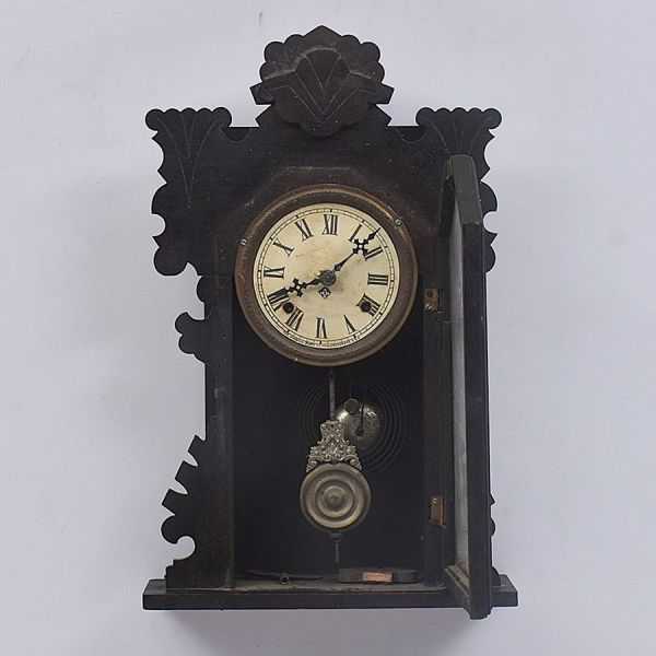 Relógio de parede `Ansonia Clock Co.` em madeira entalhada de tonalidade escura com moldura recortada em silhueta de folhagem estilizada e encimada por concha central. Porta de vidro decorada com grav
