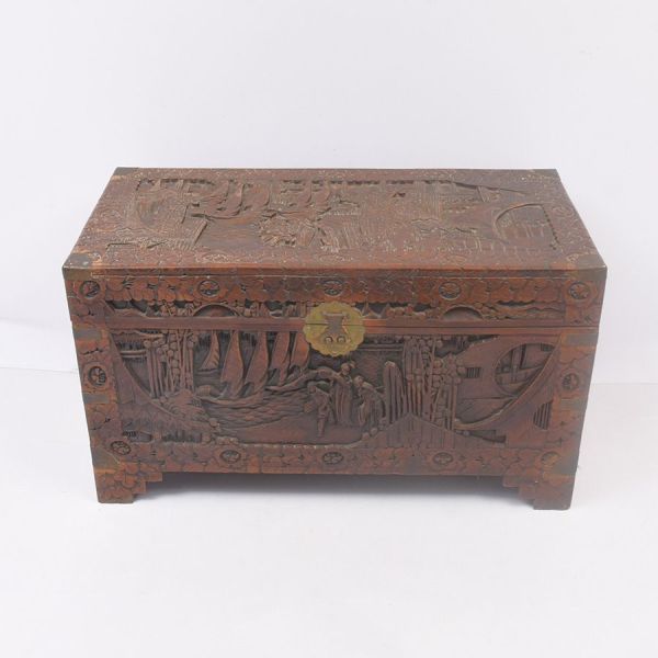 Baú em madeira de cânfora (camphor wood), chinesa, densamente entalhado em alto-relevo com cenas figurativas — pagodes, pontes, embarcações, figuras humanas e paisagens montanhosas — emolduradas por f