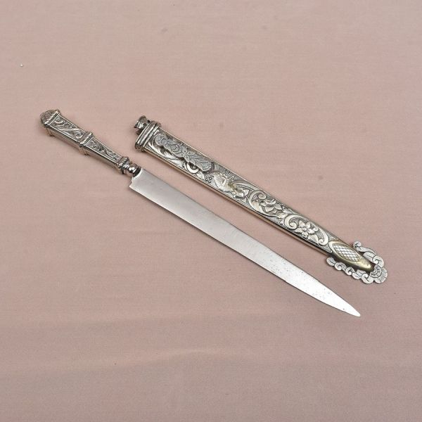 Punhal europeu em metal prateado, acompanhado de bainha integralmente decorada em relevo. O conjunto apresenta rica ornamentação de inspiração neorrenascentista, com volutas, folhagens, flores e figur