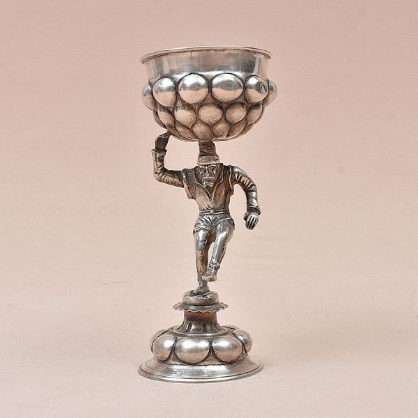 Taça figurativa em prata europeia, estilo Buckelpokal (taça de gomos). Composta por copo superior recoberto por protuberâncias esféricas em relevo disposto sobre haste representada por figura masculin