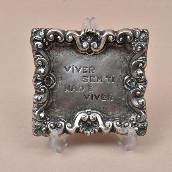 Salva quadrangular de prata portuguesa, contraste Águia com punção de garantia de teor 833 milésimos. Cinzelada com palmas e volutas e ao centro inscrição `Viver sem ti não é viver`. Med. 18 x 18 cm.