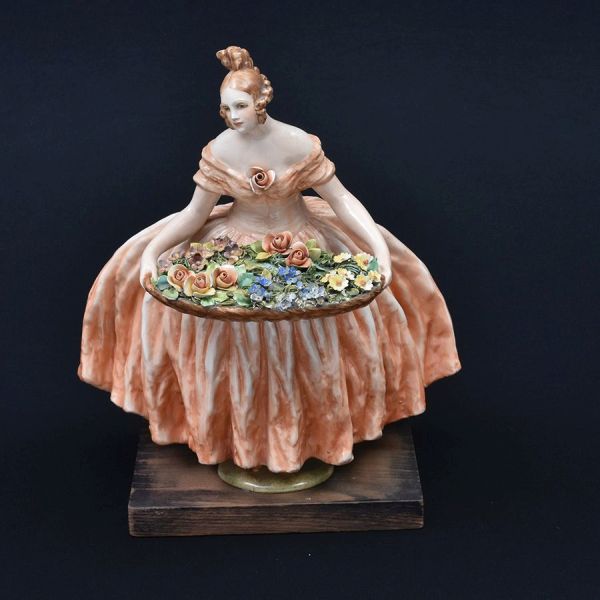 GUIDO CACCIAPUOTI (1892-1953). Escultura de porcelana italiana, manufatura "Capodimonte" em rica policromia representando dama com cesto de flores de saia volumosa e drapeada. Base de madeira. Alt. 34