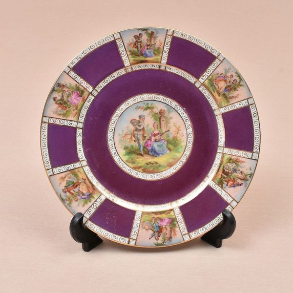 Prato de porcelana tcheca decorado em reservas com cenas de galanteio em policromia intercalados por painéis em tom de roxo com molduras em arabescos dourados. Diam. 25 cm.