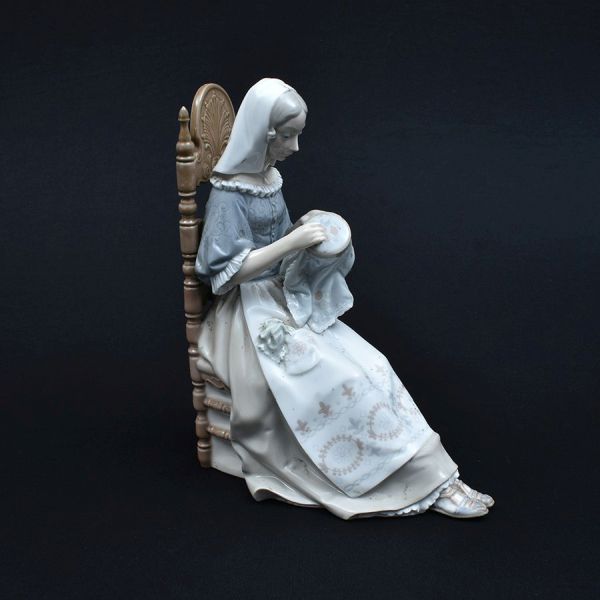 Estatueta de porcelana espanhola Lladro representando figura de mulher em atividade de costura sentada na cadeira. Alt. 28 cm.
