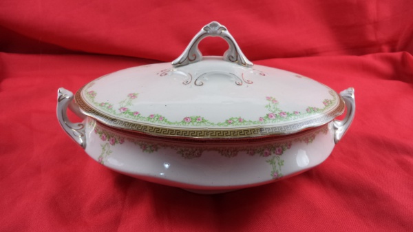 Sopeira oval com tampa e alças ornamentadas .Origem: Staffordshire, Inglaterra.Fabricante: Thos Hughes & Son (traz o carimbo oficial do fabricante com coroa e numeração "1801" em baixo-relevo na base). Época: Final do século XIX / Início do século XX (Cerca 18911910). Material:  Porcelana / Faiança inglesa fina.Decoração: Estilo Art Nouveau, decorada com delicadas guirlandas de mini rosas cor-de-rosa, ramagens verdes e bordas com friso geométrico dourado (padrão grego). Puxador e alças com realces em ouro.Uso: Ideal para servir, colecionar ou compor mesas postas de época. Medidas: 8 cm x 16 cm x 28 cm.