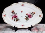 Travessa em porcelana decorada por rosas e filetação dourada em sua borda. Mede 26,5 x 42 cm. Possui bicados na borda conforme foto 2.