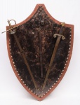 Escudo D'arma, confeccionado em madeira, forrado em couro, tecido e chapa metálica trabalhada, guarnecido com duas espadas em metal dourado e ferro e uma massa de guerra em ferro e madeira. Med:. 1,28 x 0,88.