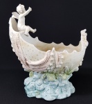 Antiga floreira europeia em porcelana biscuit policromado, realçado com gotículas duradas, representando barco com anjinho sobre o mar. Altura 22 cm, comprimento 20 cm e largura 11 cm.