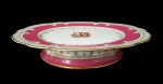 CORONEL JOÃO PINTO DA FONSECA GUIMARÃES. GRANDE STAND CAKE  EM PORCELANA COM BORDA DECORADA EM ROSE POMPADOUR REMATADA FOR FINO OURO EM GUIRLANDAS. RESERVAS COM INICIAIS ENTRELAÇADA JP. PERTENCEU AO SERVIÇO DO CORONEL JOÃO PINTO DA FONSECA GUIMARÃES. FRANÇA, MEADOS DO SEC. XIX. 26 CM DE DIAMETRO.