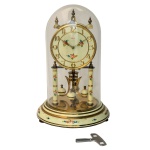 Relógio de mesa alemão com máquina aparente e mostrador circular. Estrutura em metal esmaltado de cor creme, realçada por adornos florais policromados. Pêndulo em porcelana. Redoma cilíndrica apoiada em base lisa. Marca Kundo, 400 dias. Funcionando e com chave. 30 x 20 cm