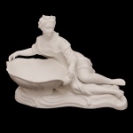 "Vide-poche" em porcelana francesa de cor branca representando figura feminina deitada sobre base, segurando bacia funda e ovalada. No verso, marca da manufatura Vion et Baury, Paris (1836-1889). 11 x 16 cm.