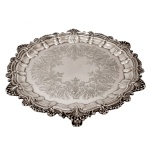 Salva em metal prateado, possivelmente inglês, com borda recortada por concheados e drapeados, centralizando larga guirlanda com folhagens e volutas. Pés curtos e recurvos. Marcas do metal e do fabricante R.B. 28 cm. 