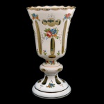 Vaso de cristal com overlay branco, ornamentado por adornos florais, frisos dourados e pequenas janelas ovais lisas. Bojo no formato de copo, apoiado em haste anelada e base circular. 25,5 x 14 cm.