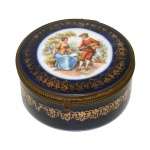 Caixa em porcelana em azul noturno. Corpo com decoração em ouro e centro da tampa com cena galante. Fecho e guarnição em metal dourado. Espelho na parte superior interna da tampa. No fundo as inscrições Made in Germany 4 x 10 cm.