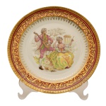 Prato em porcelana francesa Limoges com representação de cena galante ao centro. Borda com belíssima decoração em ouro e vermelha. No fundo as inscrições: Manufatura Savary, fundada em 1920. Limoges, France. 25 cm.