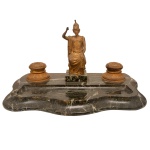 Tinteiro europeu em bronze dourado com base retangular em mármore negro rajado, com frente recortada. Apresenta par de recipientes circulares com tampas, centralizando pequena imagem de Minerva, deusa romana da Sabedoria. Séc. XIX. 24 x 42 x 26 cm.