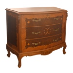 Cômoda estilo Louis XV, ornada em `marqueterie` floral, com tampo recortado em degraus. Caixa `bombée` com três gavetões superpostos, apoiada em pernas recurvas e lavradas. 98 x 118 x 56 cm. 