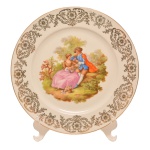 Prato em porcelana Bavária em fundo branco com dourados e representação de cena galante. Borda lisa com friso dourado. No fundo as inscrições: Manufatura Zeh Scherzer (1880-1992). Bavária, Germany. 26 cm.